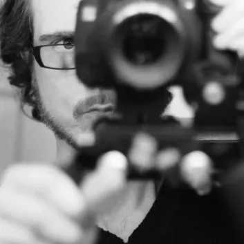 Photo en noir et blanc de Jason, qui porte des lunettes et qui pointe une caméra vers l’objectif. 