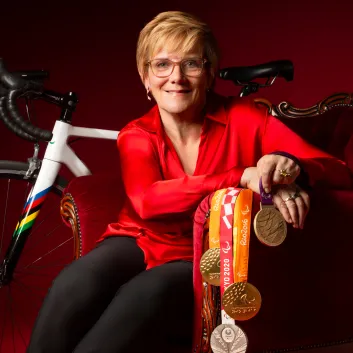 Carol porte un chemisier rouge et un pantalon noir. Elle est assise dans un fauteuil rouge devant un vélo de route et elle tient des médailles.