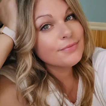 Michelle porte un t-shirt blanc. Elle a les cheveux longs, blonds et ondulés. L’un de ses bras est replié vers le haut et sa tête est appuyée sur sa main.